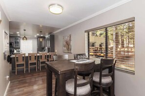 A dining area with a wooden table and chairs, a kitchen in the background, and a window with blinds. - Walk to Lake! 7bdr, Hottub, Movie Theater, Gameroom Dawn and Dusk by Avantstay (Big Bear Lake)