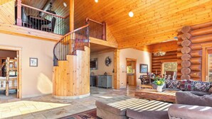 A spacious log cabin interior with a central staircase, a cozy living area with a sofa and coffee table, and a dining area with wooden furniture.