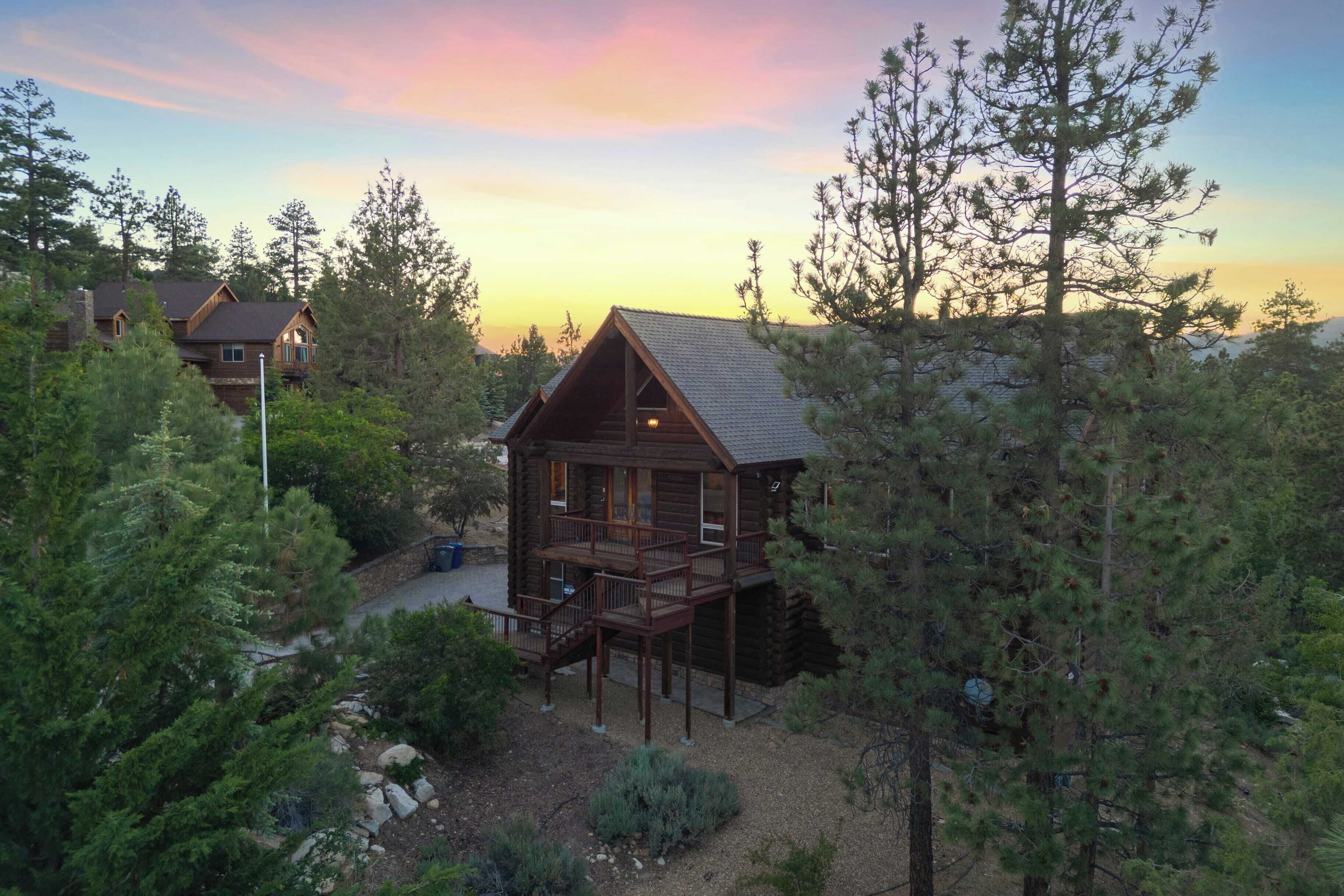 A log cabin with a deck, surrounded by dense forest and another cabin in the background.