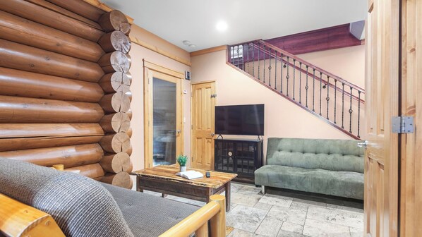 Interior de una cabaña de troncos con escalera, sofá, mesa de madera y televisor.
