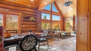 A log cabin interior with a dining area, a fireplace, and a large window offering a view of the outdoors.