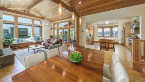 A spacious living area with a large wooden dining table, a cozy seating area with sofas, and a kitchen in the background. - The Castle Rock Estate by Avantstay Steps From Arch Cape Beach w/ Hot Tub & Sauna (Arch Cape)