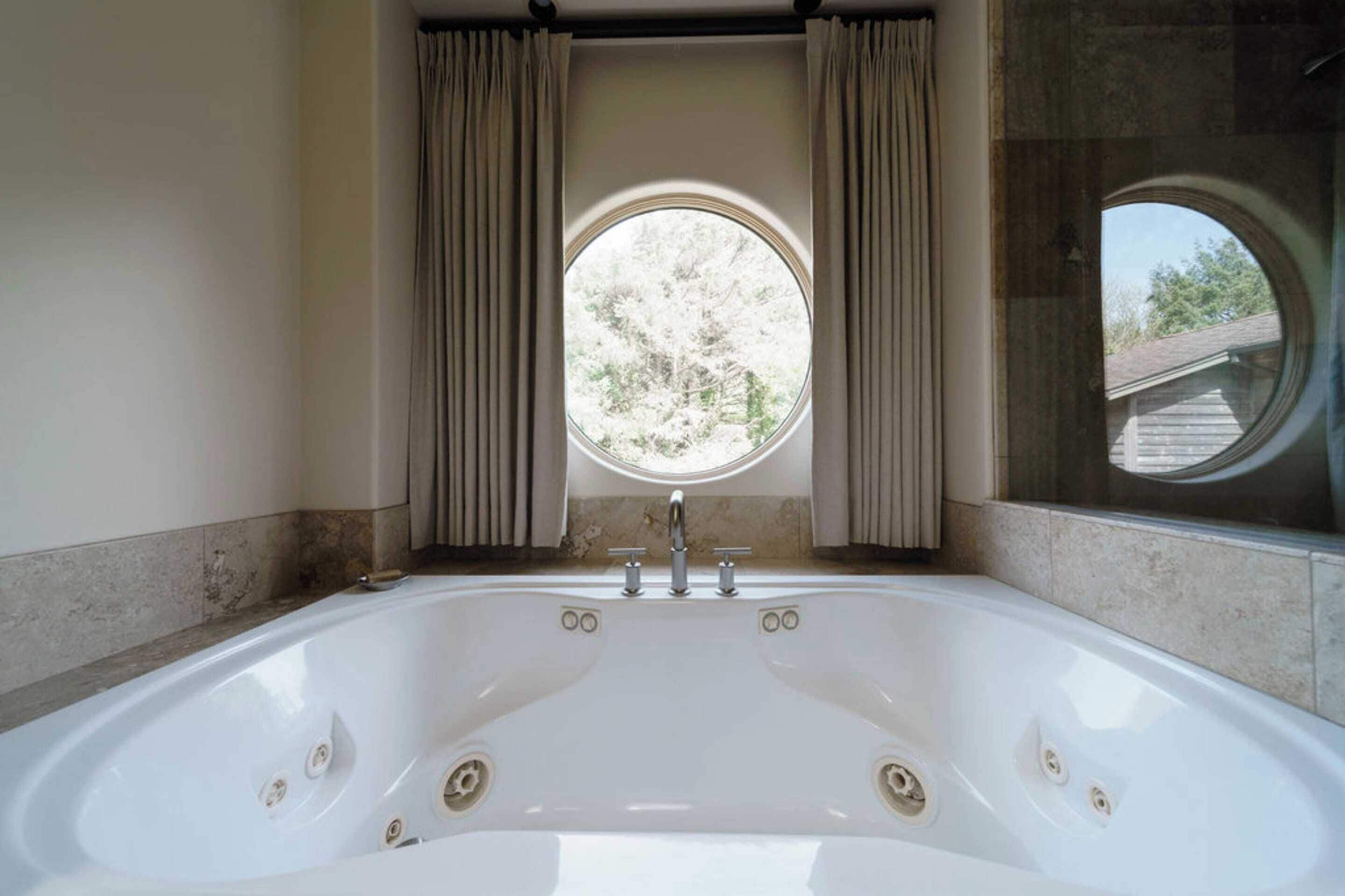 A bathroom with a large white bathtub, a window with curtains, and a view of trees outside.