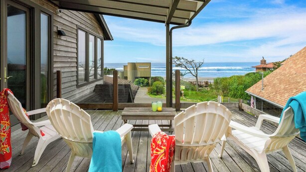 A wooden deck with white plastic chairs, a table with drinks, and a view of the ocean. - The Castle Rock Estate by Avantstay Steps From Arch Cape Beach w/ Hot Tub & Sauna (Arch Cape)