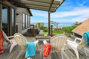 A wooden deck with white plastic chairs, a table with drinks, and a view of the ocean. - The Castle Rock Estate by Avantstay Steps From Arch Cape Beach w/ Hot Tub & Sauna (Arch Cape)