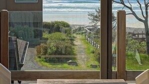 A view from a room with a large window overlooking a beach path, a wooden fence, and a stone wall. - The Castle Rock Estate by Avantstay Steps From Arch Cape Beach w/ Hot Tub & Sauna (Arch Cape)