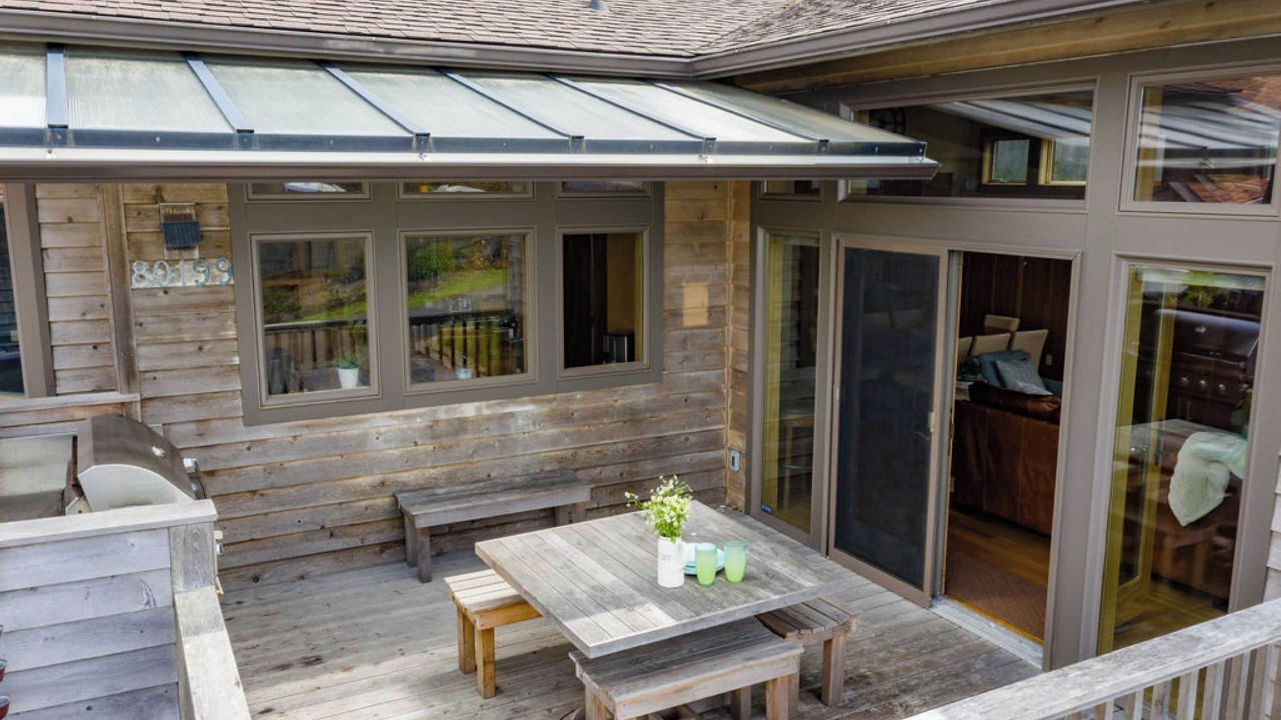 A wooden deck with a table and benches, a covered patio area, and a sliding glass door leading to an interior space.