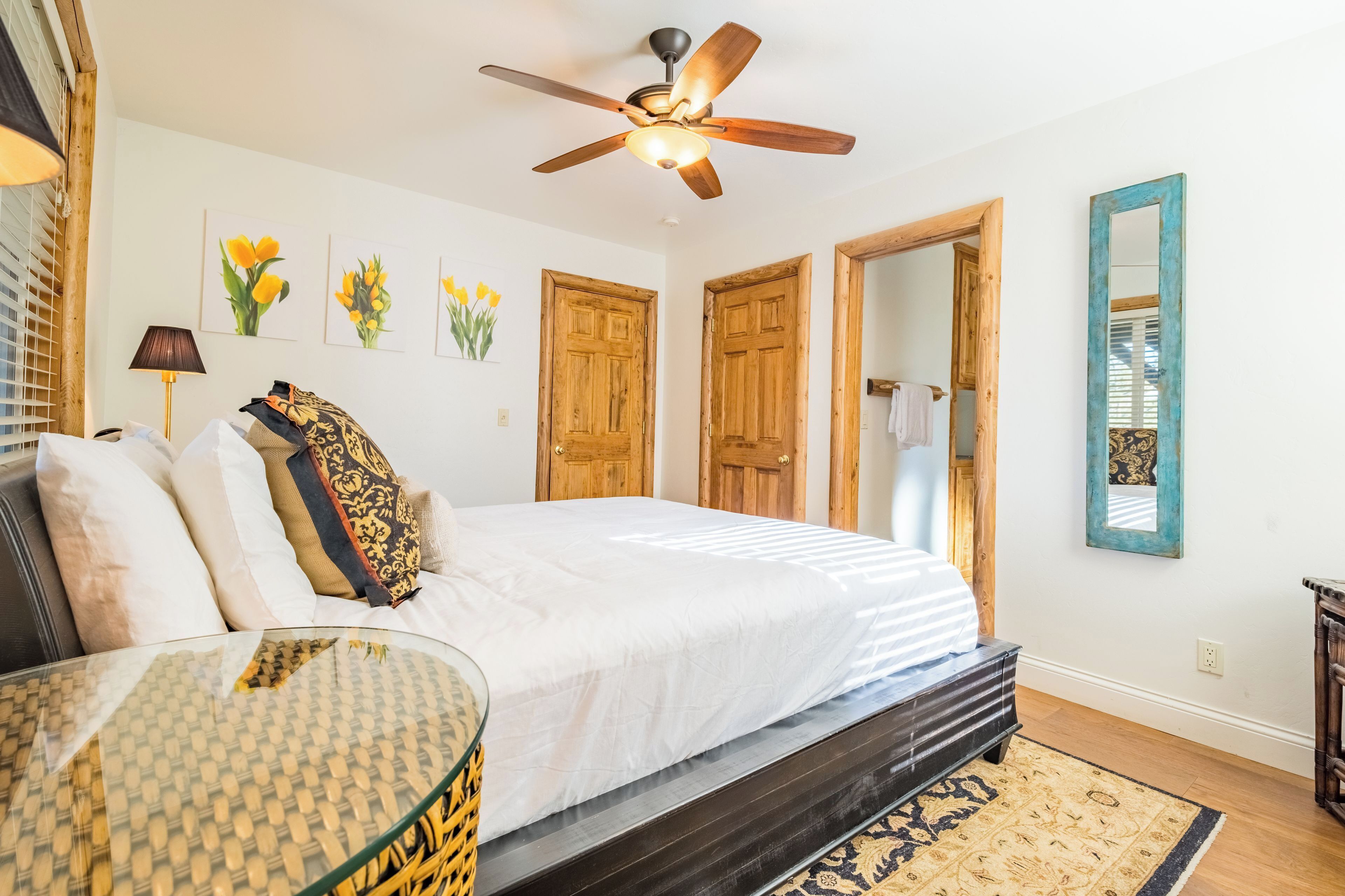 A bedroom with a bed, a glass coffee table, a ceiling fan, wooden doors, and a mirror.