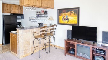 A kitchen with a refrigerator, microwave, and coffee maker, a bar area with chairs, a TV, and a speaker system.