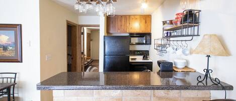 A kitchen with granite countertops, a black refrigerator, wooden cabinets, a microwave, and a hanging pot rack.