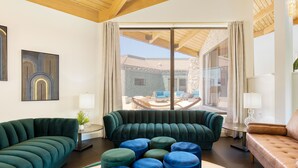 Un salon moderne avec un canapé vert, des poufs bleus et une vue sur une terrasse aménagée avec du mobilier d’extérieur.