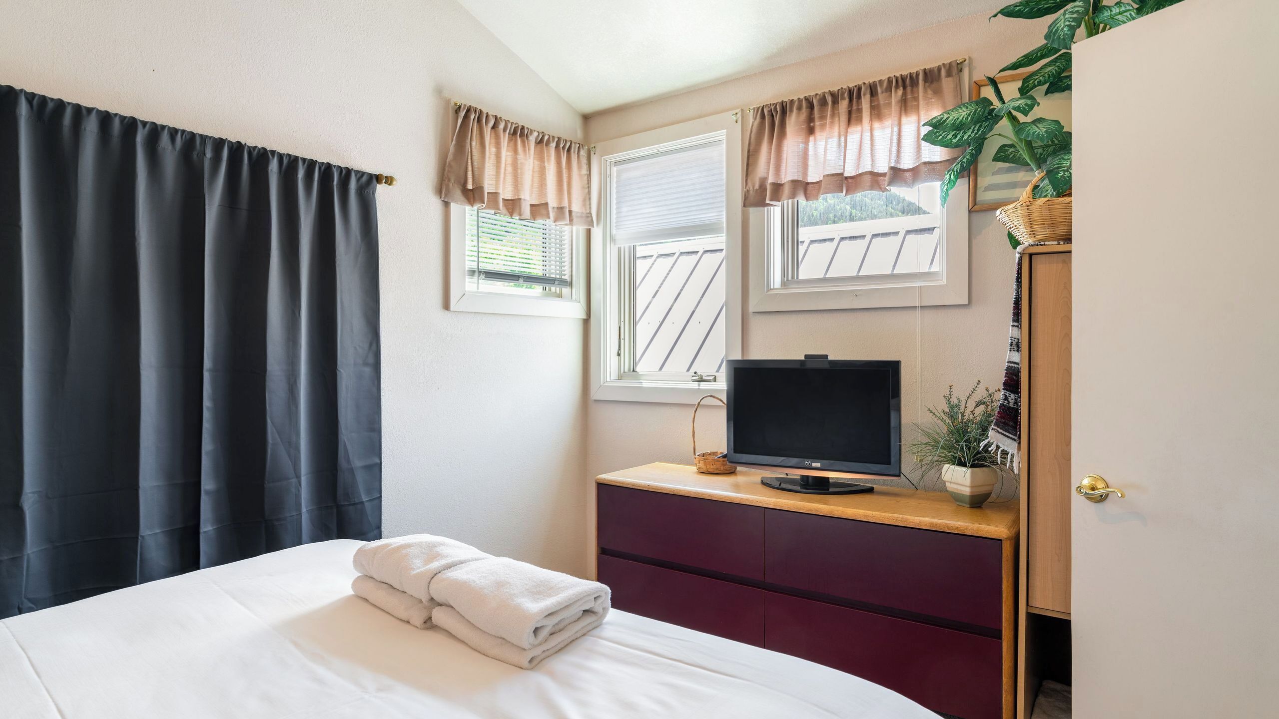 Un dormitorio con una cama, un televisor sobre una cómoda de madera, una ventana con persianas y una planta en maceta.