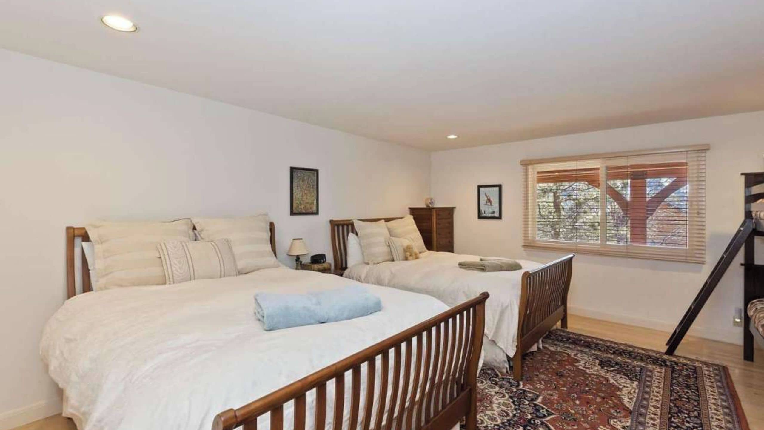 A bedroom with two beds, a wooden headboard, a nightstand, a dresser, a window with blinds, and a patterned rug.