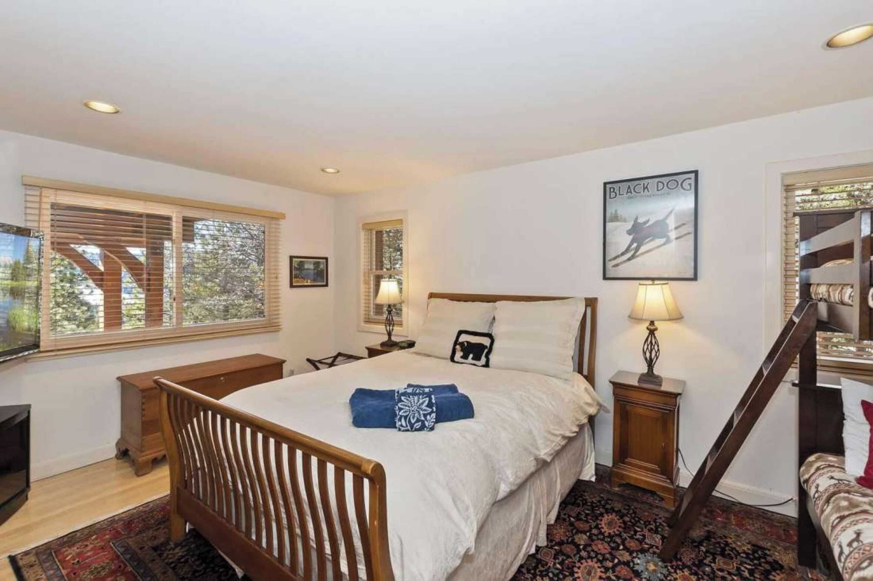 A bedroom with a large bed, two bedside tables, a wooden bench, a TV, and a window with blinds.