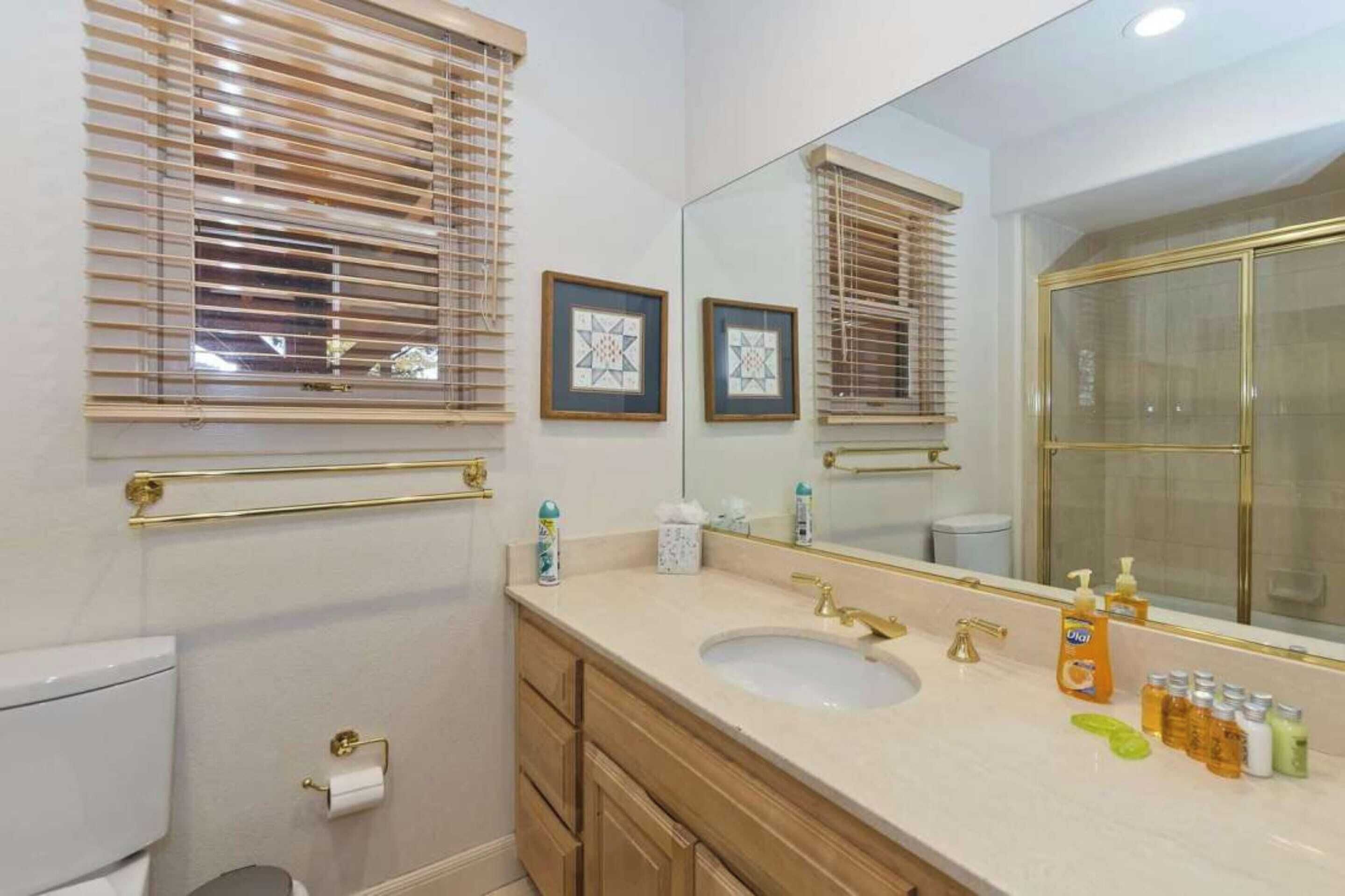 Une salle de bain avec un grand miroir, un lavabo doté de robinetterie dorée et des toilettes. Des tableaux encadrés ornent le mur, et une fenêtre est équipée de stores.