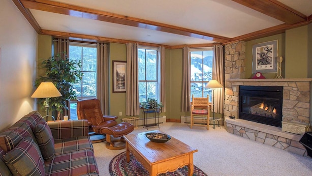 A cozy living room with a fireplace, plaid furniture, and large windows offering a view of the outdoors.