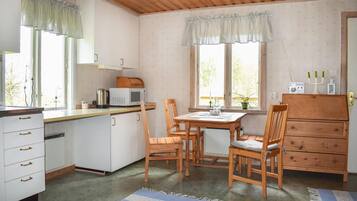 Eine kompakte Küche mit weißen Schränken, einer Mikrowelle und einer Kaffeemaschine. Es gibt einen Esstisch mit Stühlen, einen Holzschrank und ein Fenster mit Vorhängen.