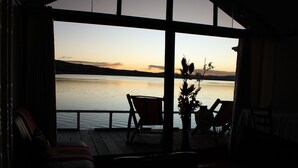 Une pièce dotée d’une grande fenêtre donnant sur un lac paisible au coucher du soleil. On y trouve deux fauteuils et une petite table sur laquelle est posé un vase rempli de fleurs.