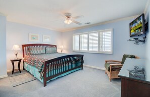 Ein Schlafzimmer mit einem Bett, zwei Nachttischlampen, einem Deckenventilator, einem Fenster mit Jalousien, einem an der Wand montierten Fernseher und einem Korbstuhl.