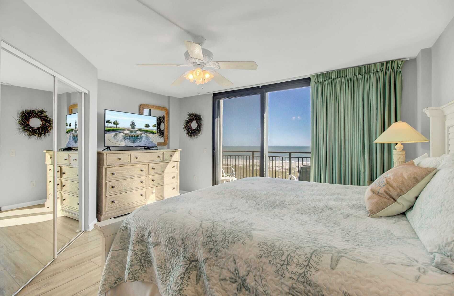 A bedroom with a bed, a dresser, a television, and a balcony with a view of the ocean.