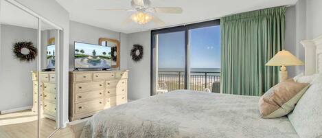 A bedroom with a bed, a dresser, a television, and a balcony with a view of the ocean.