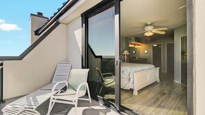A balcony with two lounge chairs, a small table, and a sliding glass door leading to a bedroom with a bed, a ceiling fan, and a nightstand.