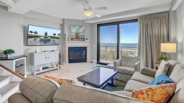 A modern living room with a fireplace, a flat-screen TV, a ceiling fan, and a balcony with a view of the ocean.