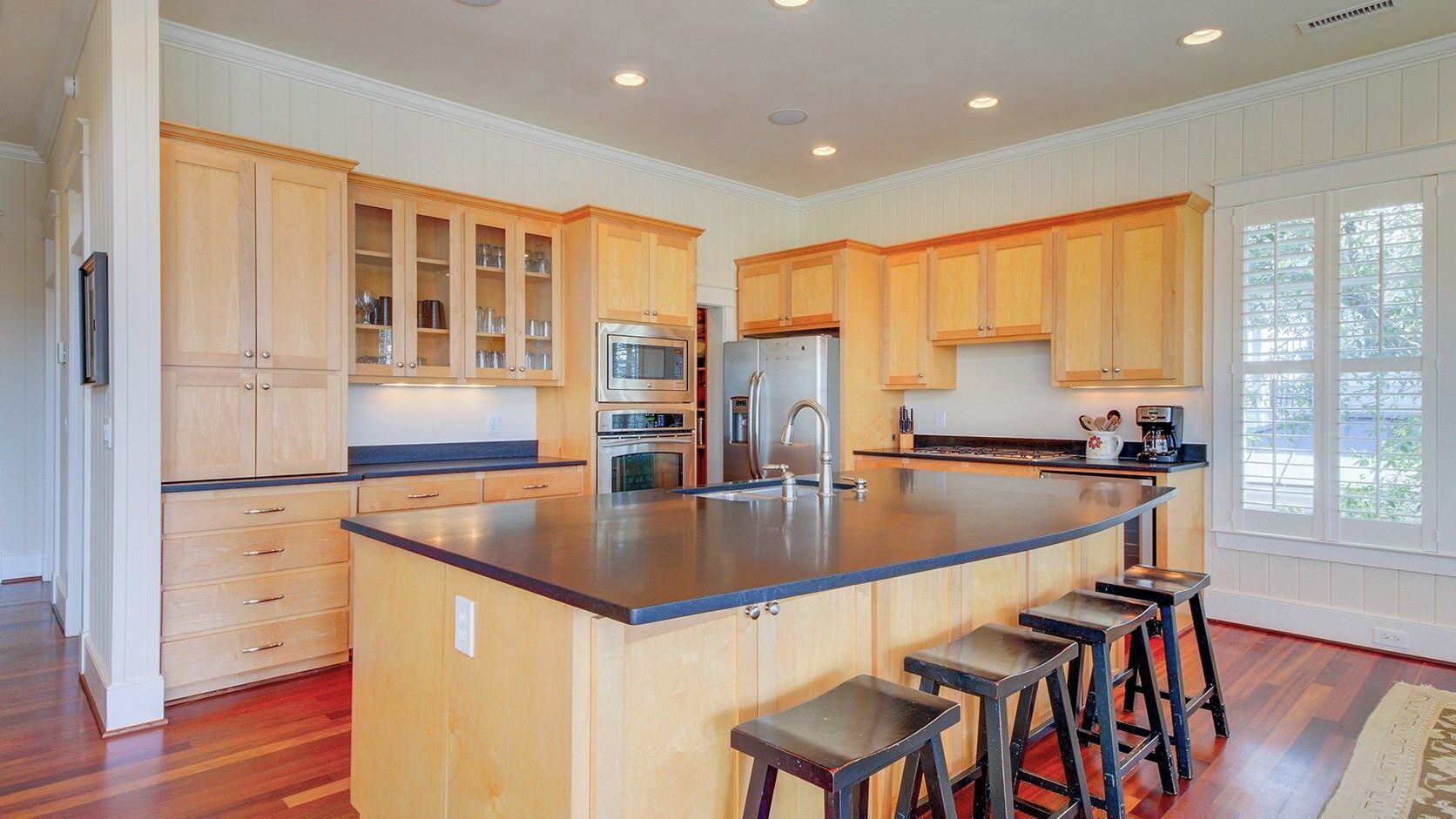 A kitchen with wooden cabinets, a central island with stools, stainless steel appliances, and a window with blinds.