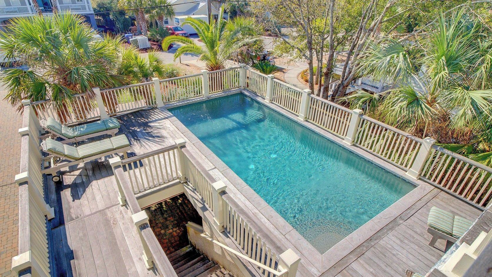A rectangular pool with a wooden deck and white railings, surrounded by palm trees and a residential area.