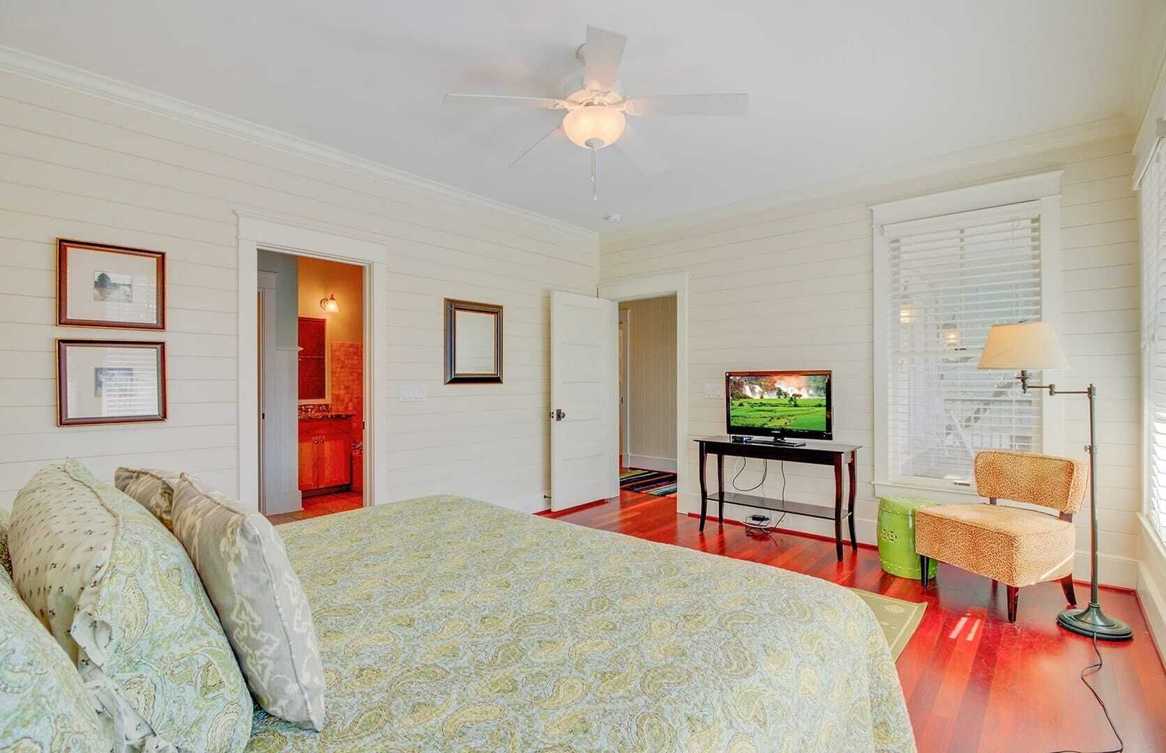 A bedroom with a bed, a television, a desk, and a chair. There is a ceiling fan and a window with blinds.