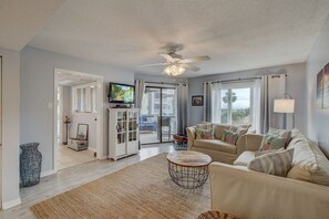 A spacious living room with a beige sofa, a television, and a ceiling fan. - 103 D Port O Call by Avantstay Close to Beach Community Pool (Isle of Palms)