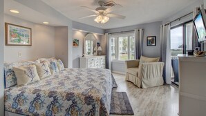 A bedroom with a large bed, a vanity, a chair, a ceiling fan, and a TV.