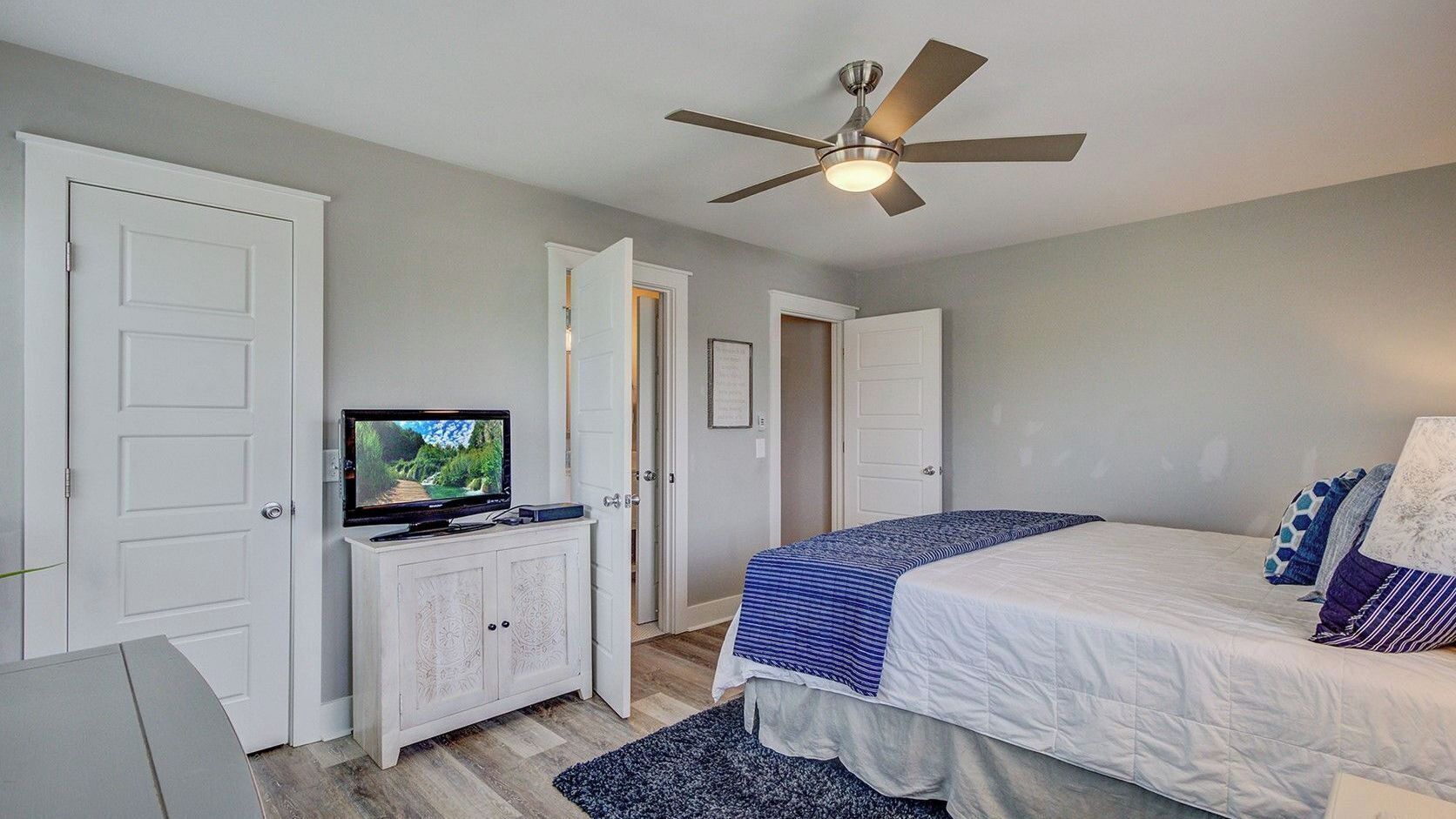 Un dormitorio con una cama, un televisor sobre un mueble blanco, un ventilador de techo y una puerta.