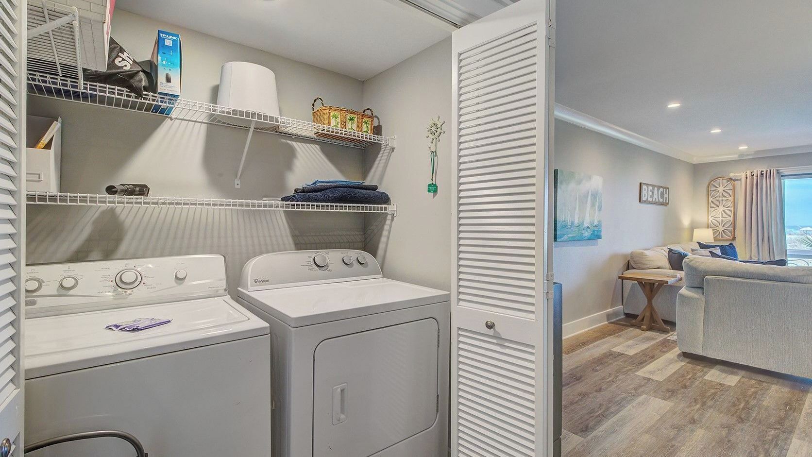 A laundry room with two washers and dryers, shelves with laundry supplies, and a view into a living area with a sofa and a table.