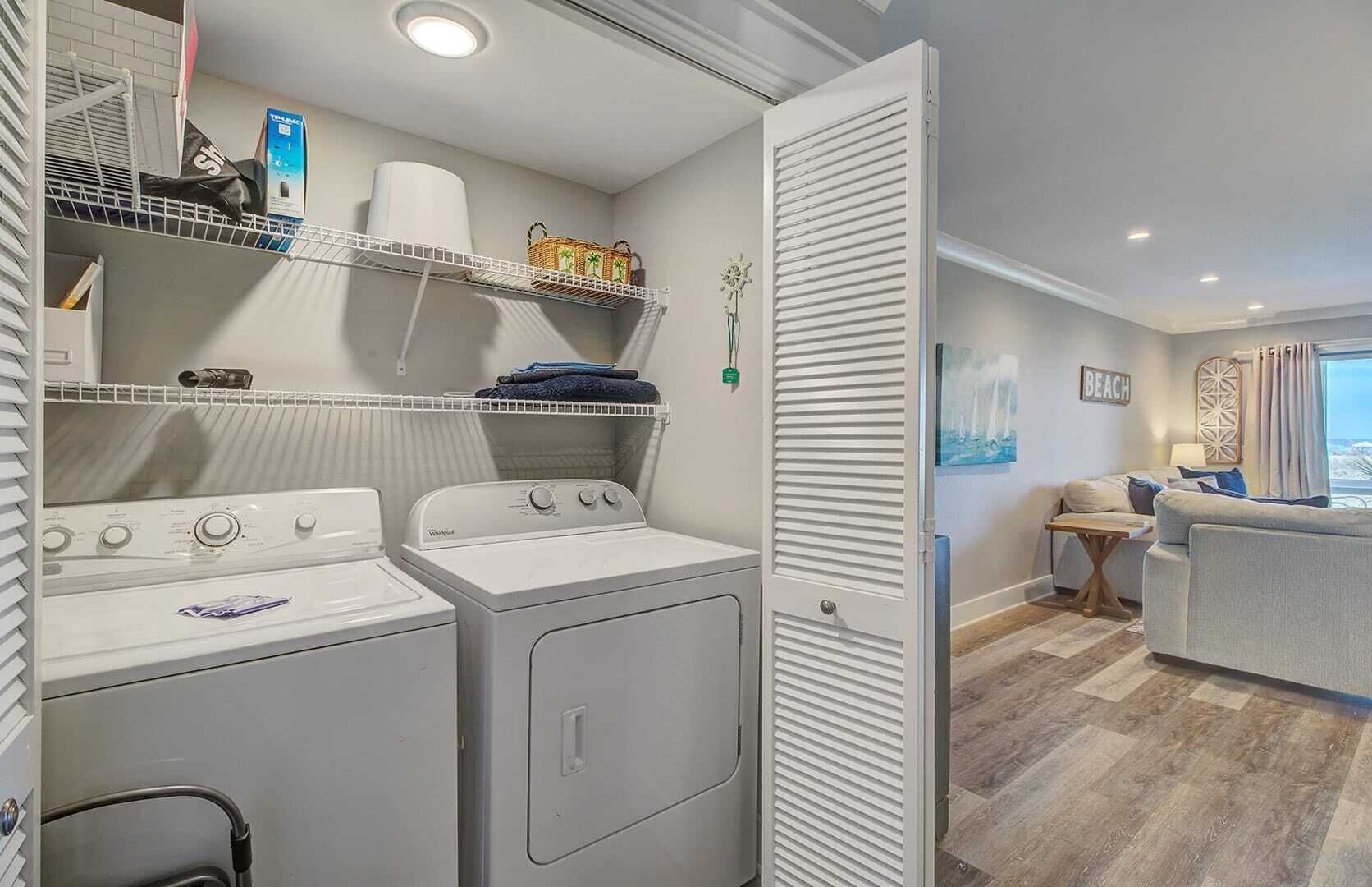 A laundry area with two washers and dryers, shelves with baskets and towels, and a view of a living room with a couch and a painting.