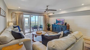 A spacious living room with a large sofa set, a coffee table, and a television. There is a sliding glass door leading to a balcony with outdoor furniture. - 11B Mariners Walk by Avantstay Direct Beach Access! Views Communal Pool (Isle of Palms)