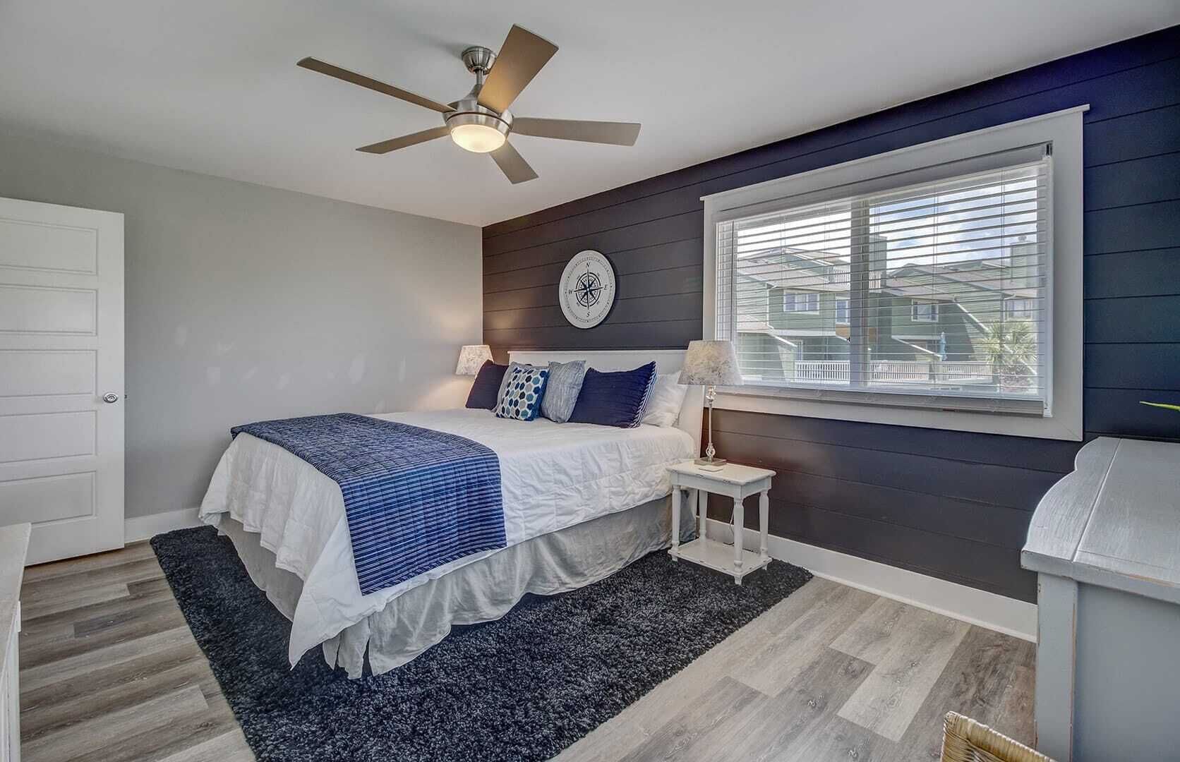 Ein Schlafzimmer mit einem Bett, einem Nachttisch, einem Deckenventilator, einem Fenster mit Jalousien und einer Wanduhr.