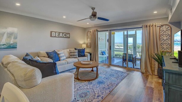 A spacious living room with a sofa, coffee table, and a ceiling fan. A large window offers a view of the beach. - 11B Mariners Walk by Avantstay Direct Beach Access! Views Communal Pool (Isle of Palms)