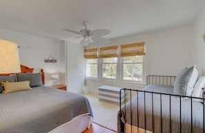 Une chambre avec deux lits, un ventilateur de plafond, une fenêtre avec des stores et une table de chevet avec une lampe.