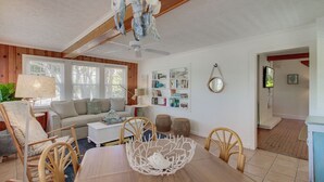 Ein gemütliches Wohnzimmer mit Holzbalkendecke, einer Couch, einem Couchtisch und einem Esstisch mit Stühlen. Ein Bücherregal mit Büchern und Dekorationselementen sowie ein kleiner runder Spiegel an der Wand sind ebenfalls vorhanden.
