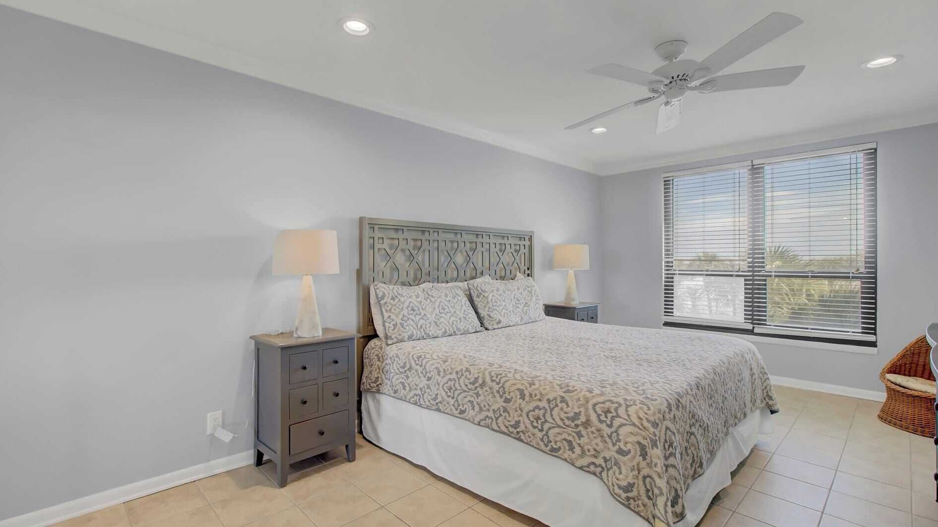 A bedroom with a bed, two bedside tables with lamps, a ceiling fan, and a large window with blinds.