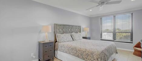 A bedroom with a bed, two bedside tables with lamps, a ceiling fan, and a large window with blinds.