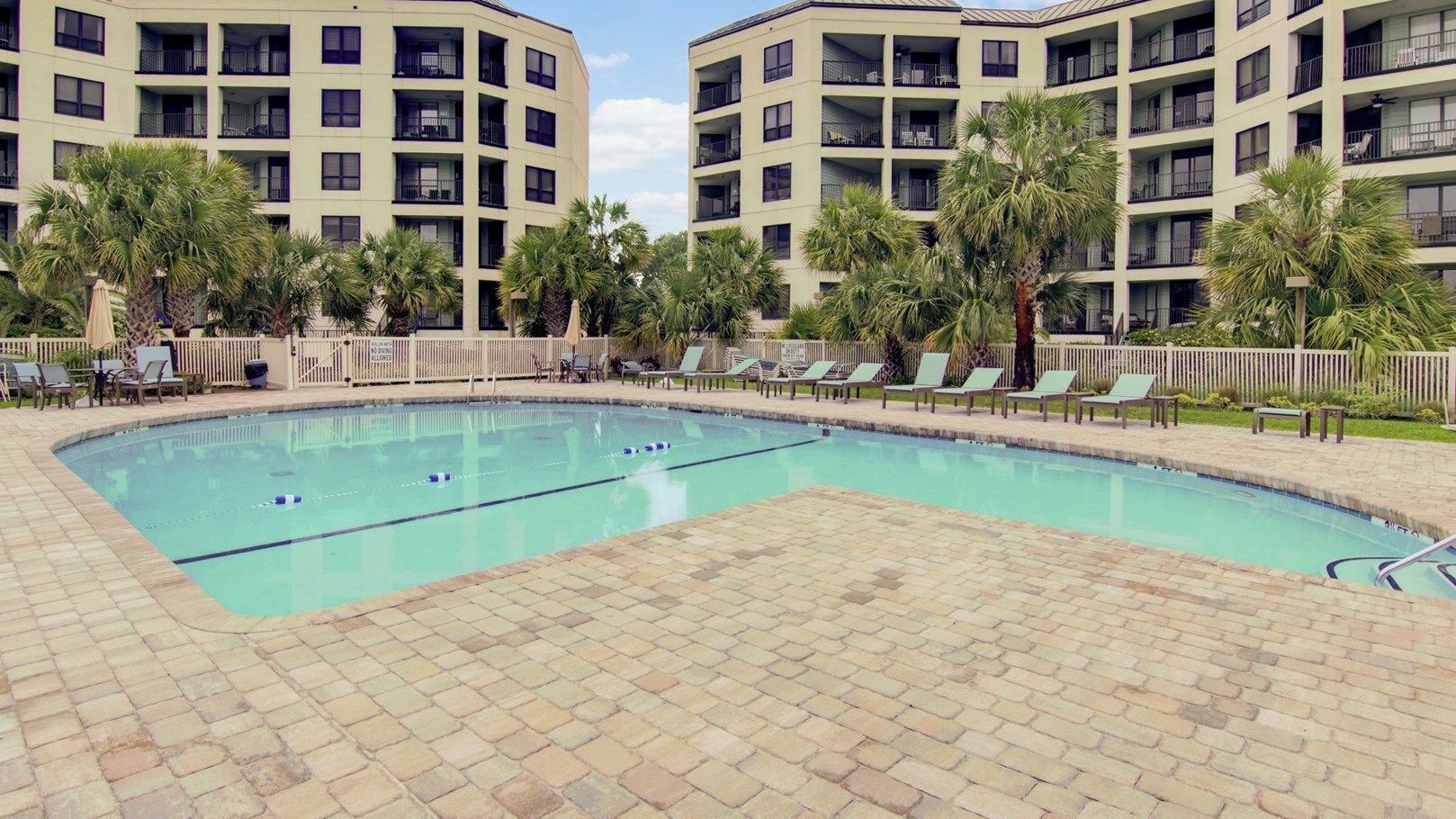 A swimming pool with lounge chairs and palm trees, surrounded by multi-story buildings.