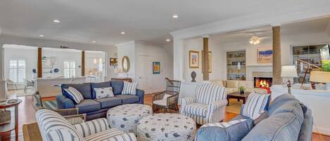 A spacious living room with a blue sofa set, striped armchairs, and ottomans, a fireplace, and a kitchen in the background.