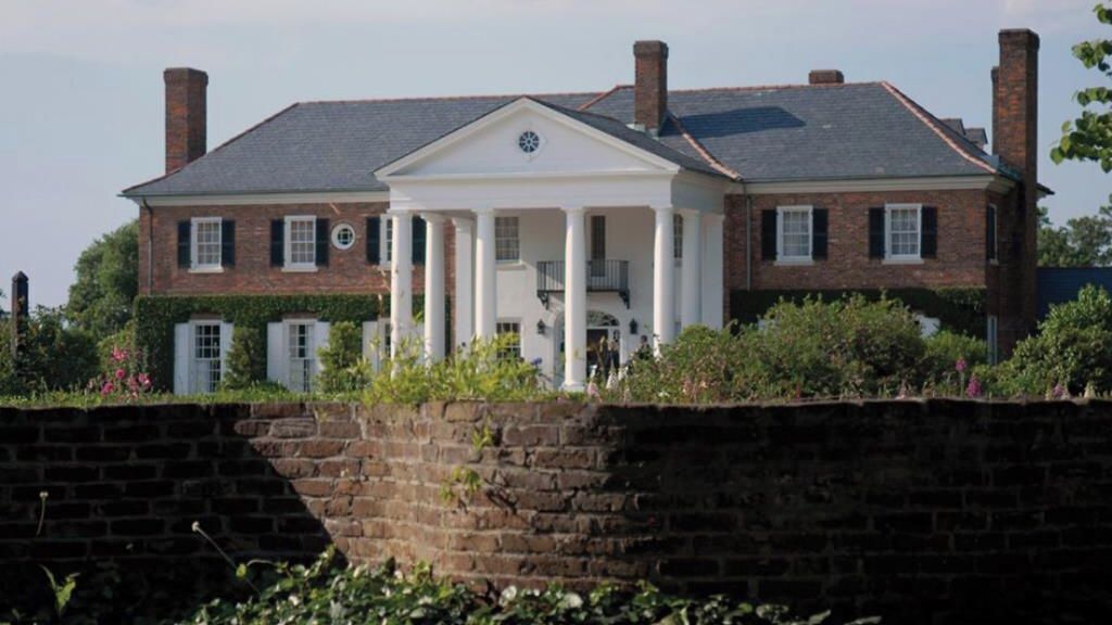 A large brick building with a central white portico, surrounded by greenery and a stone wall.