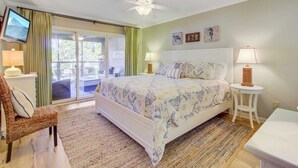 A bedroom with a bed, a television, a chair, a table, and a ceiling fan. - 405 Yacht Harbor by Avantstay Marina Views + Pool Access + Outdoor Lounge Near Golf & Beach (Isle of Palms)