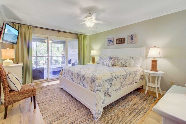 A bedroom with a bed, a television, a chair, a table, and a ceiling fan. - 405 Yacht Harbor by Avantstay Marina Views + Pool Access + Outdoor Lounge Near Golf & Beach (Isle of Palms)