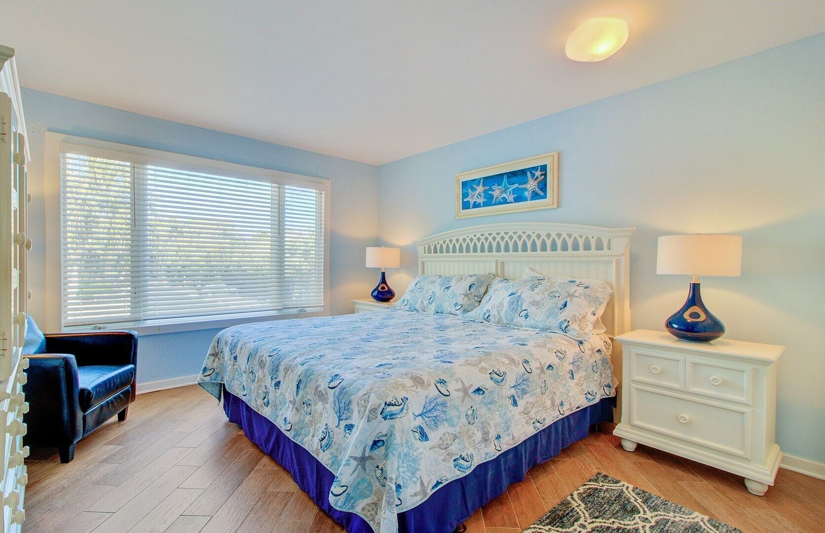 Un dormitorio con cama, dos mesitas de noche, una lámpara de noche, una ventana con persianas, una silla y una alfombra.