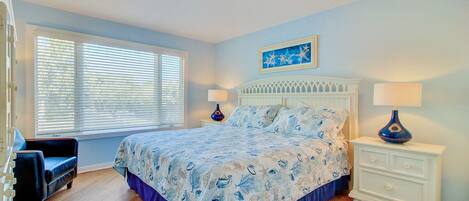 Un dormitorio con cama, dos mesitas de noche, una lámpara de noche, una ventana con persianas, una silla y una alfombra.