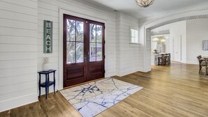 A spacious hallway with wooden floors, a large wooden door, and a view into the kitchen area.